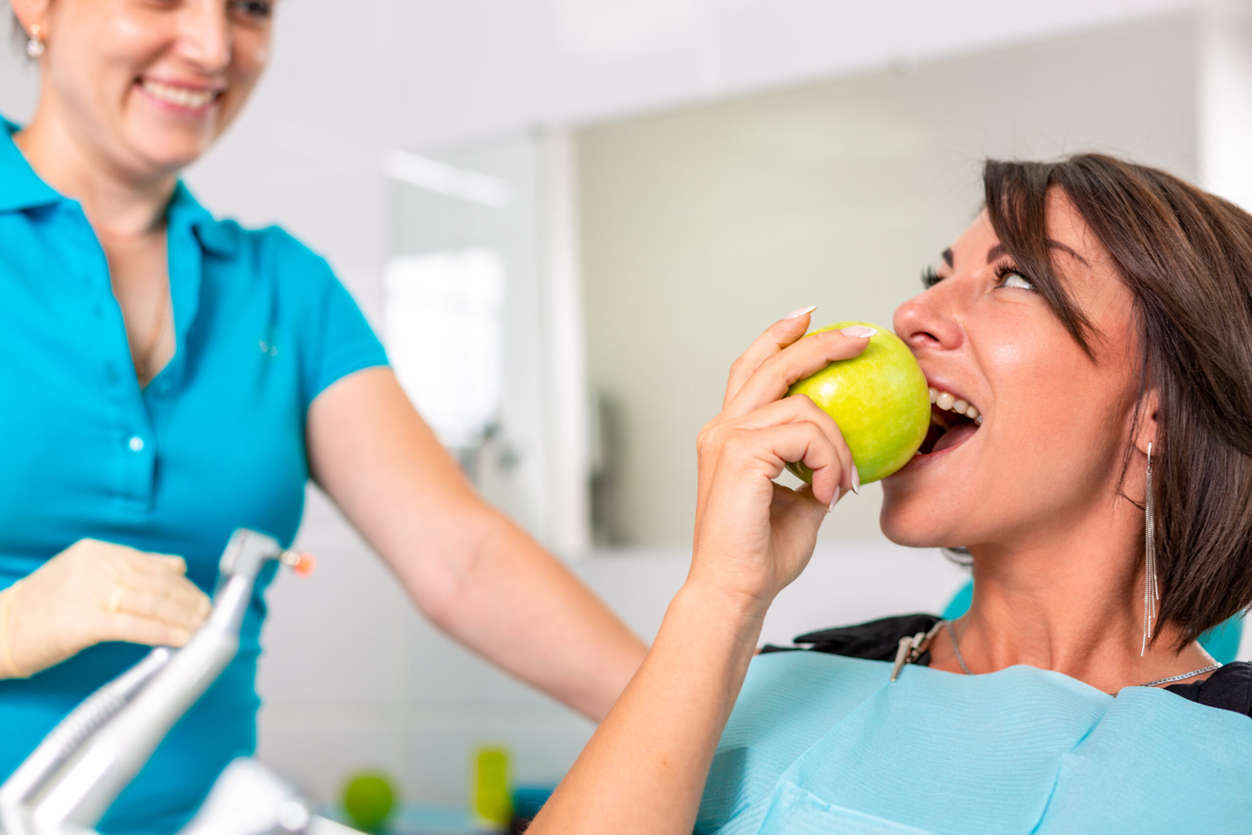 Eine Patientin bei der Zahnbehandlung, die einen grünen Apfel isst, während eine Zahnarzthelferin lächelt.