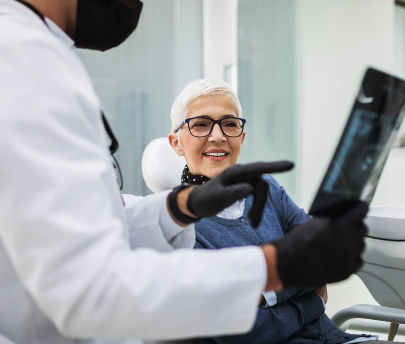 Ältere Frau mit kurzen, grauen Haaren und Brille, die einem Zahnarzt bei einer Untersuchung zusieht.
