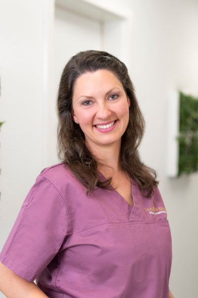 Frau in lila Uniform mit lächelndem Gesicht in einem modernen Zahnarztpraxis-Umfeld.