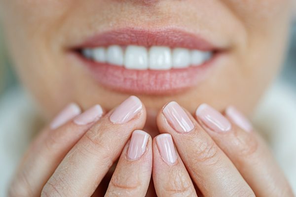 Nahaufnahme von Händen einer Person, die sanft das Gesicht hält, mit gepflegten, rosafarbenen Nägeln und einem Lächeln im Hintergrund.